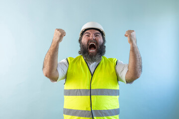 euphoric adult man engineer with beard and wearing helmet and safety vest, with raised fists,...