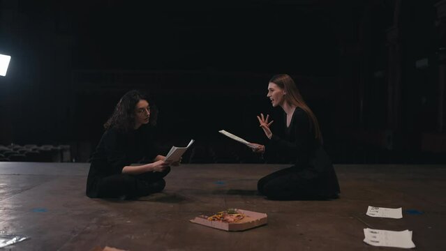 Confident two female actresses in black suits sit on stage holding papers with a script in their hands and expressively expressing their ideas and acting out roles during a rehearsal for a play in an