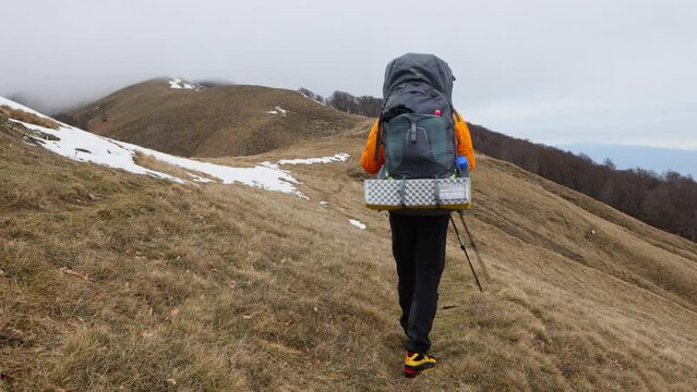 A lone hiker trekking through snowy patches in a mountainous landscape under a cloudy sky during winter.