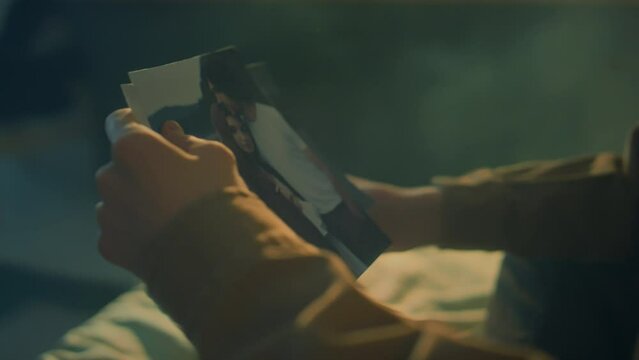 Side tilt footage of young Caucasian man sitting in bed at dawn looking at old photos of his friends or ex girlfriend and getting angry, throwing photos away