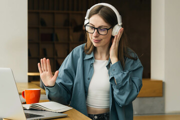A woman with glasses and headphones sits in front of a laptop and waves hello.