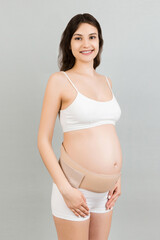 Portrait of pregnant woman in underwear wearing pregnancy belt at gray background with copy space....