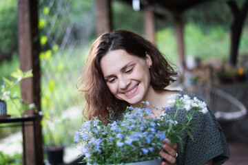 Young happy smiling Caucasian woman holding pot of flowers while sitting on terrace of country cottage, summer lifestyle portrait