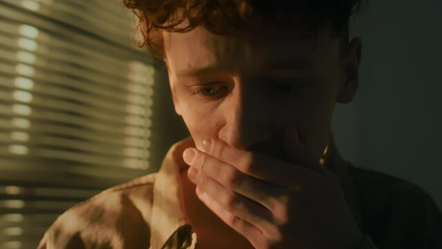 Tilt closeup of young Caucasian man feeling desperate and broken after reading obituary message on phone in dark room at dawn