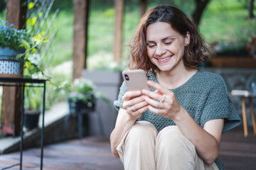 Young happy smiling Caucasian woman sitting on wooden terrace with looking at smartphone using online app