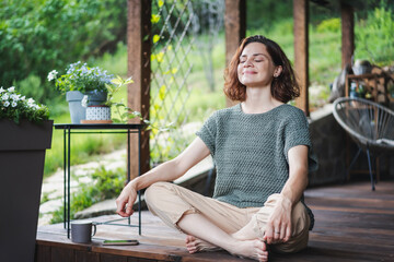 Young happy smiling Caucasian woman sitting on wooden terrace in lotus position, enjoying summer