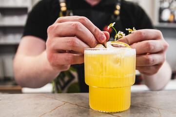 Close-up of hands garnishing a yellow cocktail with citrus and small flowers.