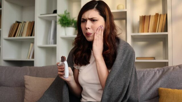 Portrait of young brunette woman eating chocolate bar and having toothache sitting on couch at home Sad female feeling pain in tooth because caries gum disease or sensitive teeth indoors
