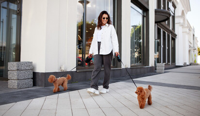 Stylish woman walks down the city street with two small dogs on leashes. Urban lifestyle with cute...