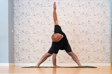 Senior man in black outfit practicing a triangle pose yoga on a mat in a bright, spacious room with...