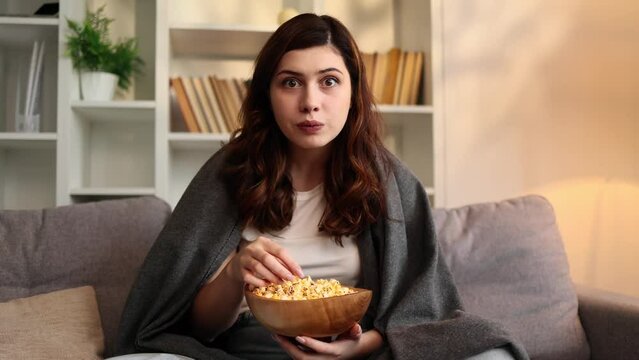 Close up portrait of addicted focused young woman looking at camera while watching interesting TV program film movie and eating popcorn at home Enchanted brunette girl cannot take her eyes off indoors