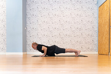 Senior man in black outfit practicing a low plank yoga pose on a mat in a bright, spacious room with a white brick wall and wooden floor. 