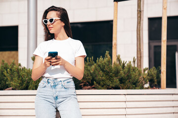 Young beautiful smiling hipster woman in trendy summer clothes. Carefree model posing in street at sunset. Positive female. Holds smartphone, uses phone apps, looks at telephone screen