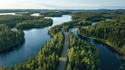 Aerial view of a winding road cutting through lush green woods, flanked by serene blue lakes under the summer sun in Finland, highlighting the natural beauty.