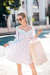 Young beautiful smiling hipster woman in trendy summer clothes. Sexy carefree model in the street at sunset. Positive female holding shopping bag. Posing outdoors. In sunglasses