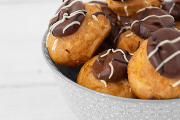 Chocolate profiteroles stacked in a decorative bowl. On a white wood background
