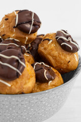 Chocolate profiteroles stacked in a decorative bowl. On a white wood background