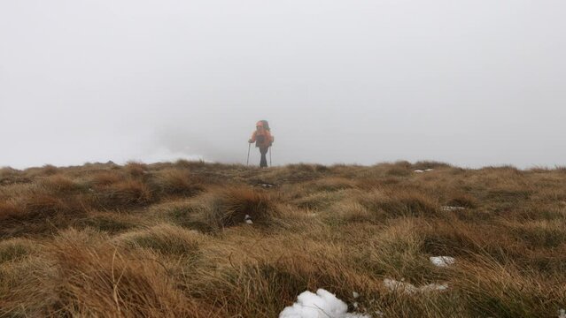 A lone hiker trekking through snowy patches in a mountainous landscape under a cloudy sky during winter.