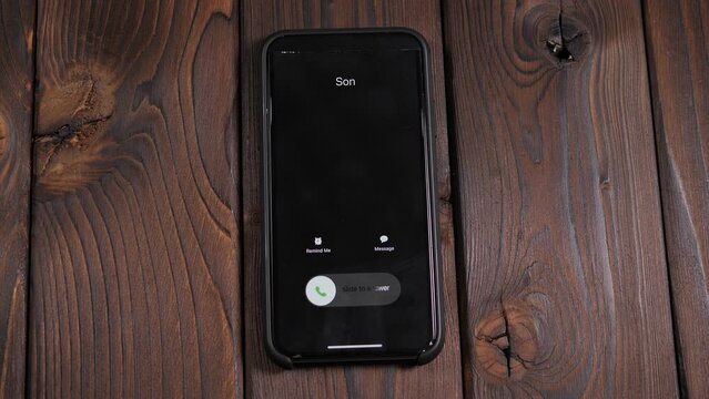 A close-up of an incoming call from his son on a smartphone screen on a wooden background, a man answers the call.