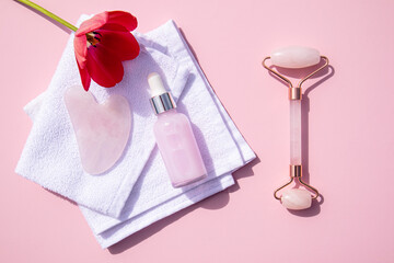 Roller massager, gua sha scraper, natural massage oil in a bottle with a dropper on a white towel. pink background. Health and beauty concept