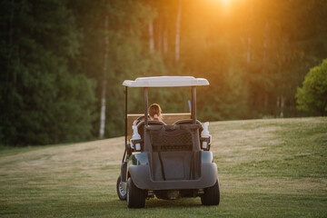  A young girl drives a golf cart on a grassy field at sunset, with a backdrop of lush green trees. © Raivo