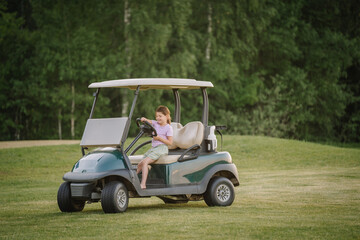 A young girl sits in a golf cart on a grassy field, smiling and holding the steering wheel, with a background of lush green trees.