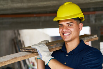 Portrait of smiling young male carpenter wearing workwear carrying planks on shoulder at...