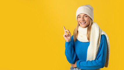 A cheerful woman wearing a white knit hat, scarf, and blue sweater points upwards with her finger....