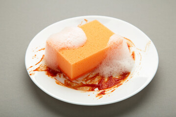 White round dirty plate with red sauce, orange foam sponge cleaning dish on grey background.