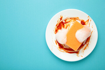 White round dirty plate with red sauce, orange foam sponge cleaning dish on blue background.