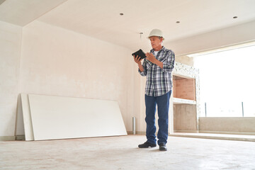 Full length of senior male architect wearing hardhat using tablet PC inside incomplete house