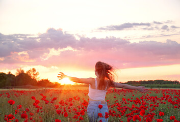Beautiful young girl running in flower poppy field at sunset. Landscape with lots of red flowers in meadow, natural idyllic spring sunset