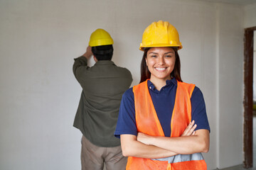 Portrait of smiling young female construction worker in vest standing with arms crossed against coworker at incomplete house