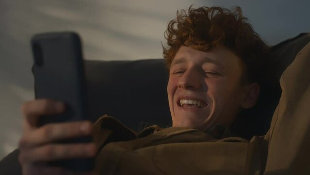 Chest up footage of ginger haired gen Z guy laughing hard at joke while surfing net on phone in bed