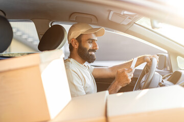 Indian man wearing a hat is behind the wheel of a car filled with packages, checking his phone for...
