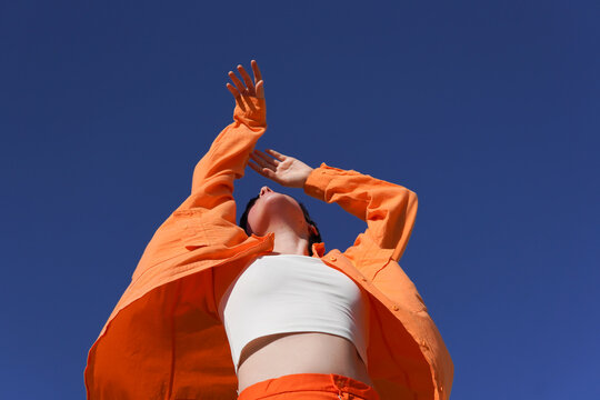 Woman in orange casuals dancing with arms raised under clear blue sky on sunny day