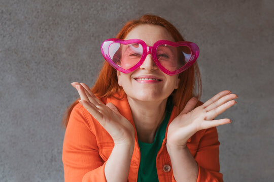 Happy woman wearing heart shape eyeglasses in front of gray wall