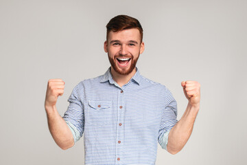 Cheerful guy showing his biceps, gesturing hands up and smiling on grey studio background, copy...