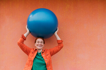 Happy woman holding fitness ball on head in front of orange wall