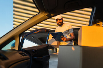 Indian delivery worker wearing a cap and t-shirt is unloading cardboard boxes from a car at a...