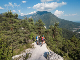 Fototapeta premium Young couple enjoying spectacular scenery, smiling while sitting on electric mountain bikes and giving a high five to each other.