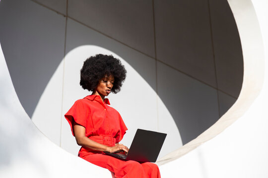 Woman in red jumpsuit with Afro hairstyle using laptop sitting on white wall