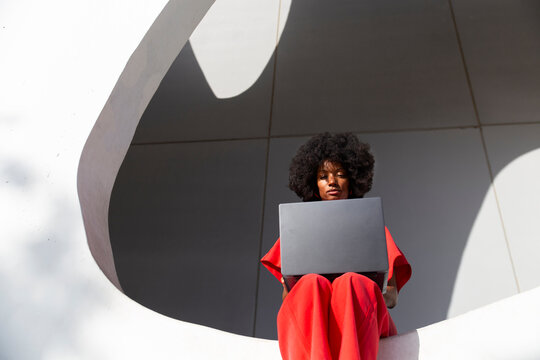 Woman with Afro hairstyle using laptop sitting on white wall