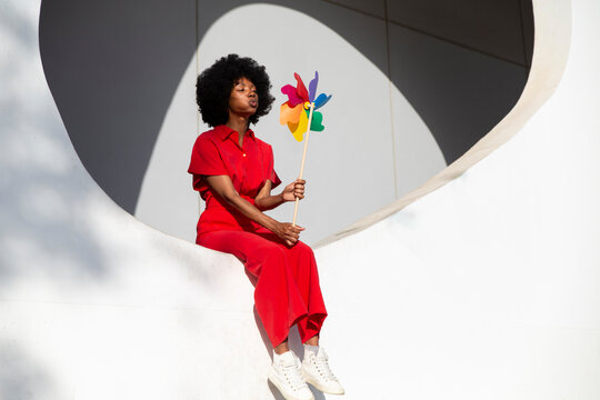Mature woman in red jumpsuit blowing at colorful pinwheel toy sitting on white wall