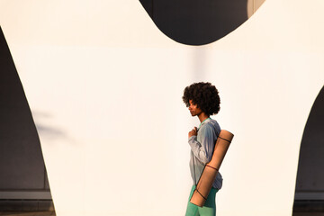 Woman with Afro hairstyle carrying exercise mat walking by white wall