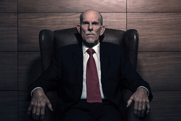 Senior businessman in suit and red tie sits in chair in front of a wall.