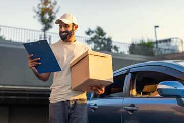 Indian delivery driver is standing next to a blue car in a sunny parking lot. He is holding a large...