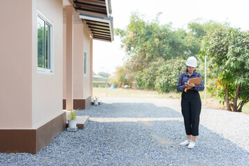 Asian female inspector in safety helmet writing on clipboard, standing near house. Represents...