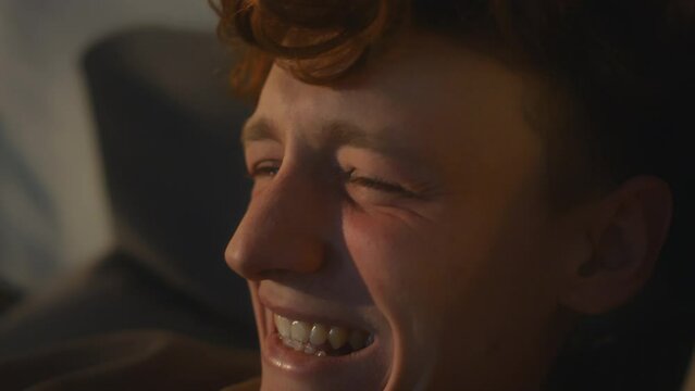 Side closeup of Caucasian male zoomer laughing so hard and closing eyes while watching sitcom or joking online with friends indoors