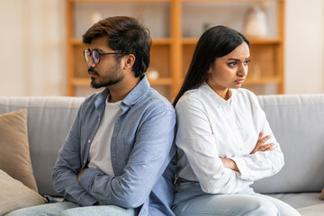Indian young couple sits back-to-back on a couch, both with arms crossed, appearing to be in the...